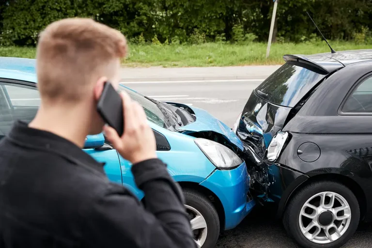 A driver calling for help on a smartphone at the scene of a rear-end collision between a blue hatchback and a black car, illustrating steps to take after a car accident and personal injury legal advice.