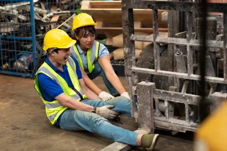 A warehouse worker in a yellow hard hat and safety vest sitting on the floor in pain after a forklift accident, with a colleague assisting, ideal for workers' compensation claims and workplace injury legal services.