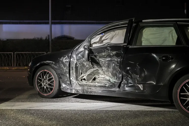 Side-view of a black car with severe T-bone collision damage at night, showing deployed side-curtain airbags and crushed passenger doors for a car accident lawsuit or insurance claim.