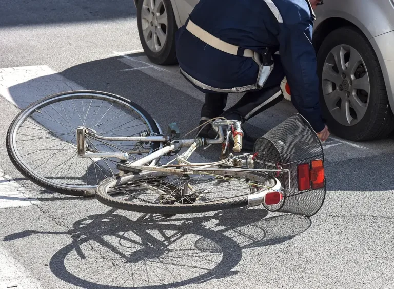 A bicycle with a front basket lying on a crosswalk after a collision, with a police officer documenting the accident scene involving a silver car.