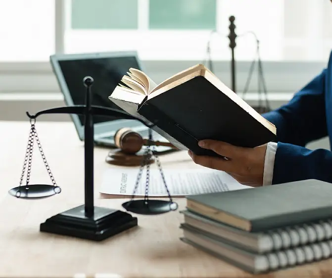 An attorney in a blue suit holding an open law book at a desk with a laptop, gavel, and scales of justice, illustrating detailed legal research and case preparation.