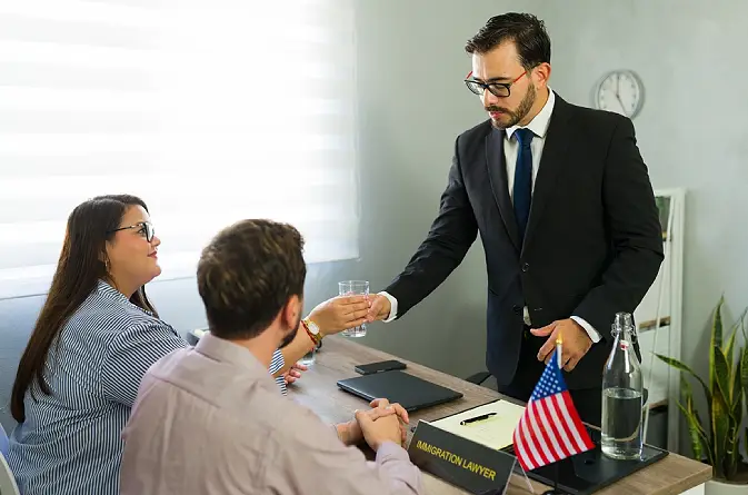 An immigration lawyer in a suit handing a glass of water to a couple during a legal consultation in a modern office with an American flag and professional desk signage.
