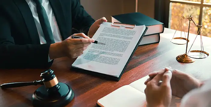 Close-up of a lawyer in a suit pointing to a legal contract on a clipboard during a consultation, with a gavel and scales of justice on a wooden desk.